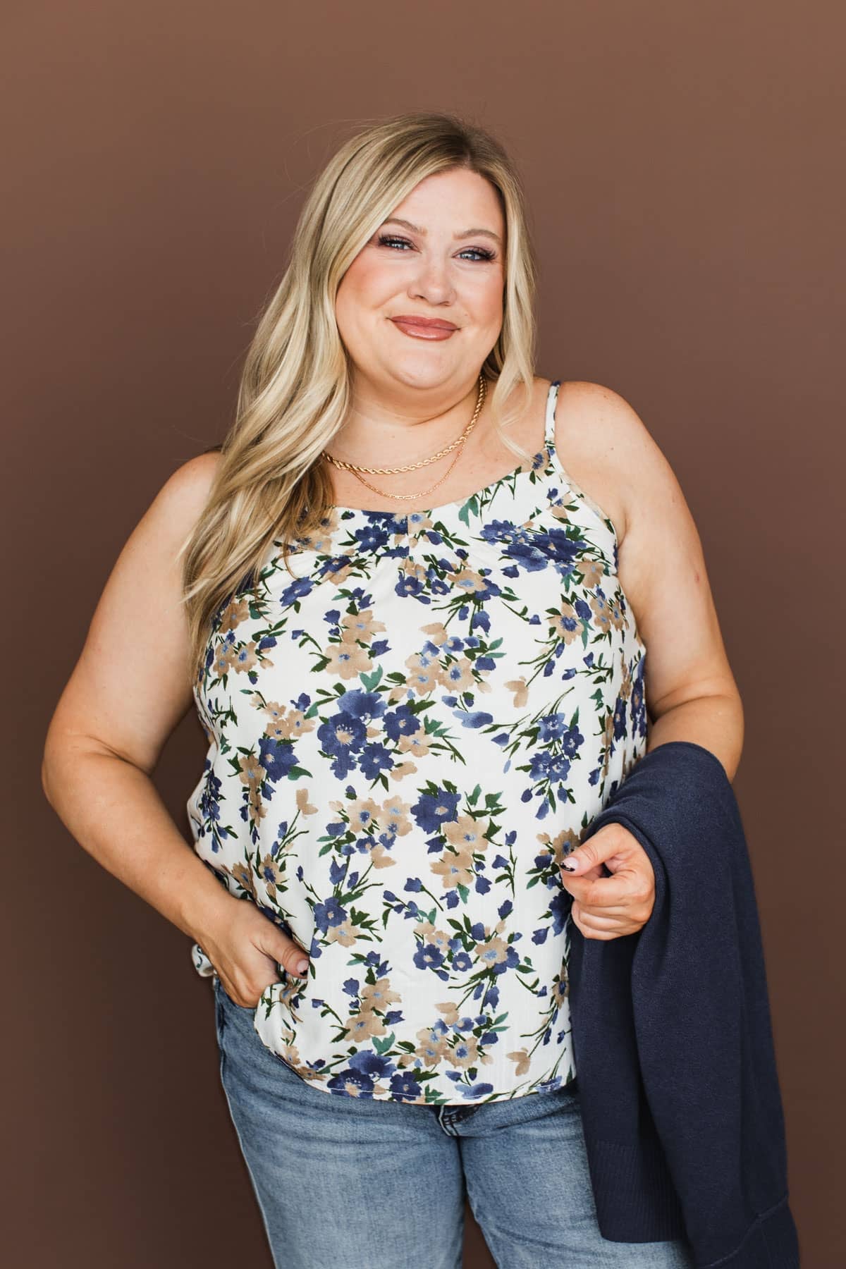 A woman modeling a sleeveless white camisole top with a blue and beige floral pattern, paired with blue jeans and holding a navy cardigan against a solid brown studio background.