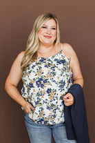 A woman modeling a sleeveless white camisole top with a blue and beige floral pattern, paired with blue jeans and holding a navy cardigan against a solid brown studio background.
