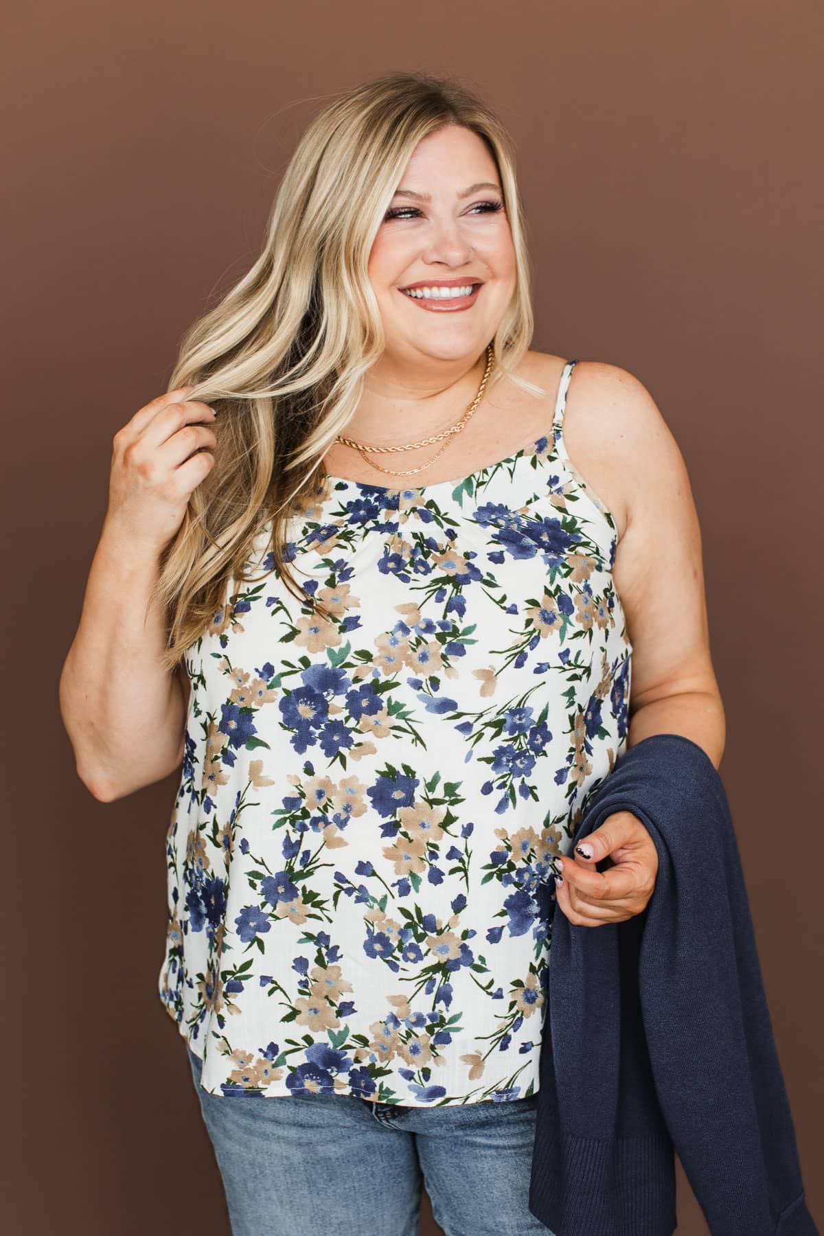 A smiling blonde woman wearing a white spaghetti-strap tank top with a blue and beige floral pattern, paired with blue jeans and holding a navy blue cardigan against a solid brown studio background.