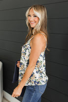 A woman wearing a sleeveless floral camisole top with blue and beige flowers on a white background, paired with blue jeans, standing against a dark gray shiplap wall.