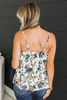 A woman viewed from behind wearing a sleeveless floral camisole top with thin straps and blue jeans, standing against a dark gray shiplap wall.