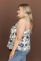 A woman shown from the side wearing a sleeveless floral camisole top with blue and tan flowers on an off-white background, paired with blue jeans, against a solid brown studio backdrop.