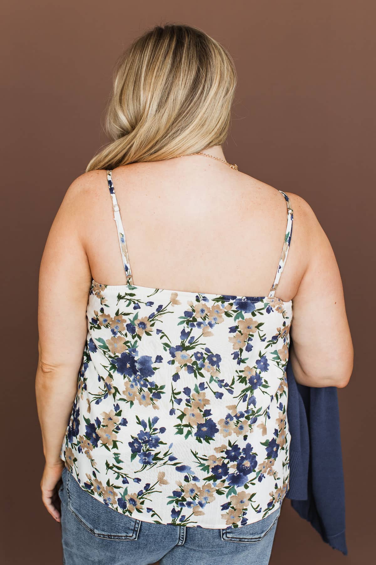 A woman shown from the back wearing a white spaghetti-strap camisole top with a blue and beige floral pattern, paired with blue jeans, against a solid brown background.
