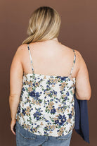 A woman shown from the back wearing a white spaghetti-strap camisole top with a blue and beige floral pattern, paired with blue jeans, against a solid brown background.